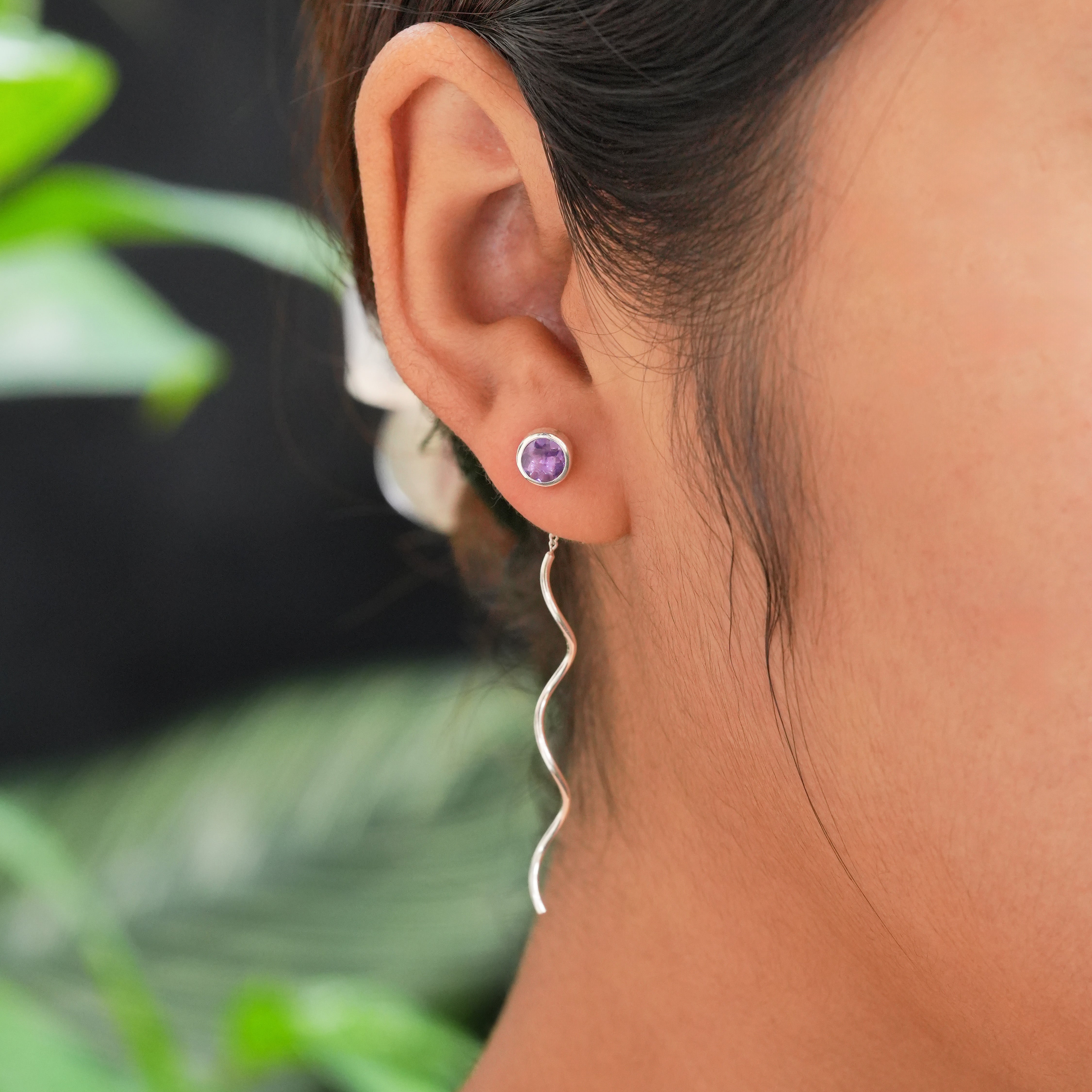 Faceted amethyst stud earrings with flowing curled sterling silver wire drops worn on an ear for size reference