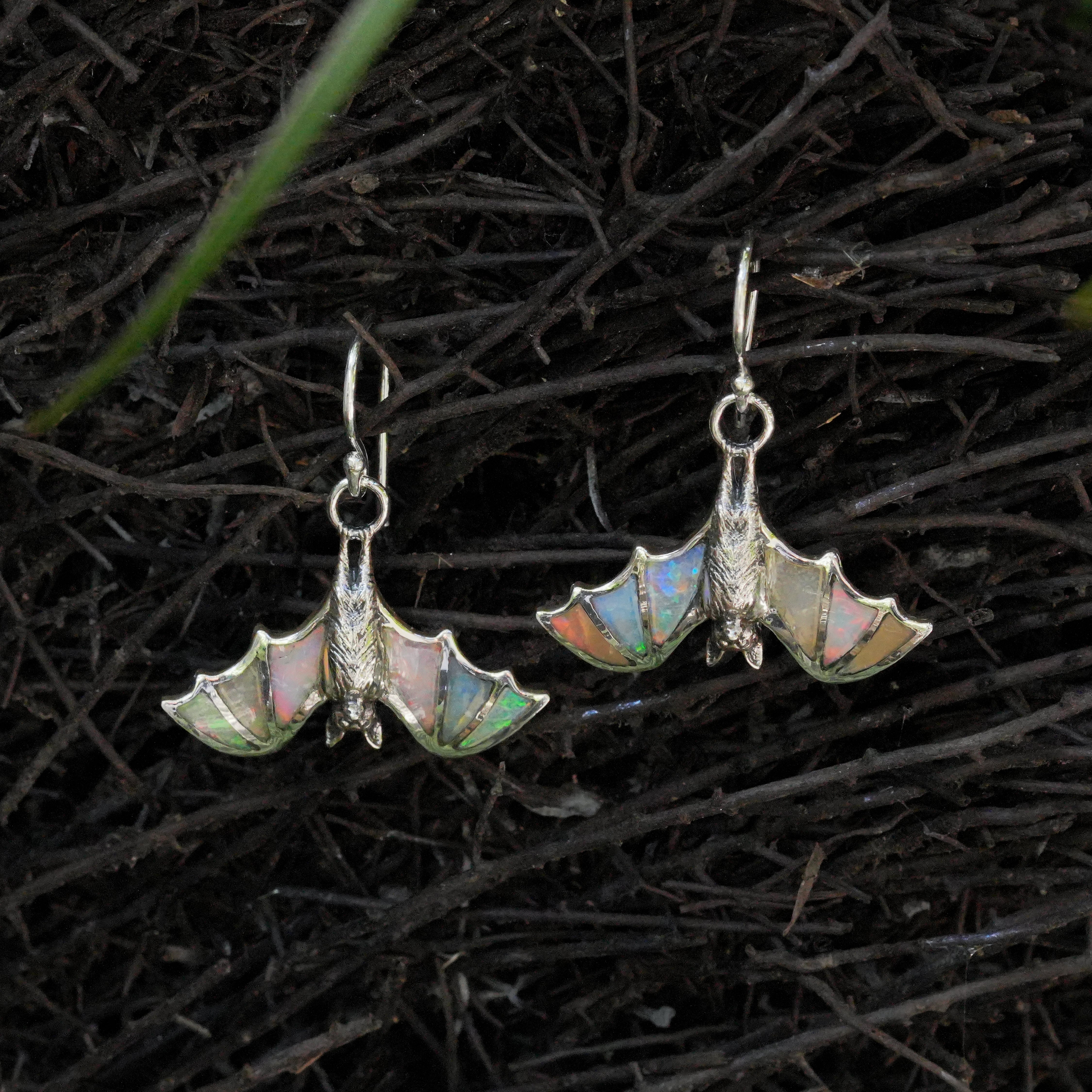 Angled view of hanging bat earrings with Ethiopian opal inlay arranged to resemble bat wing veining, showing natural play‑of‑color
