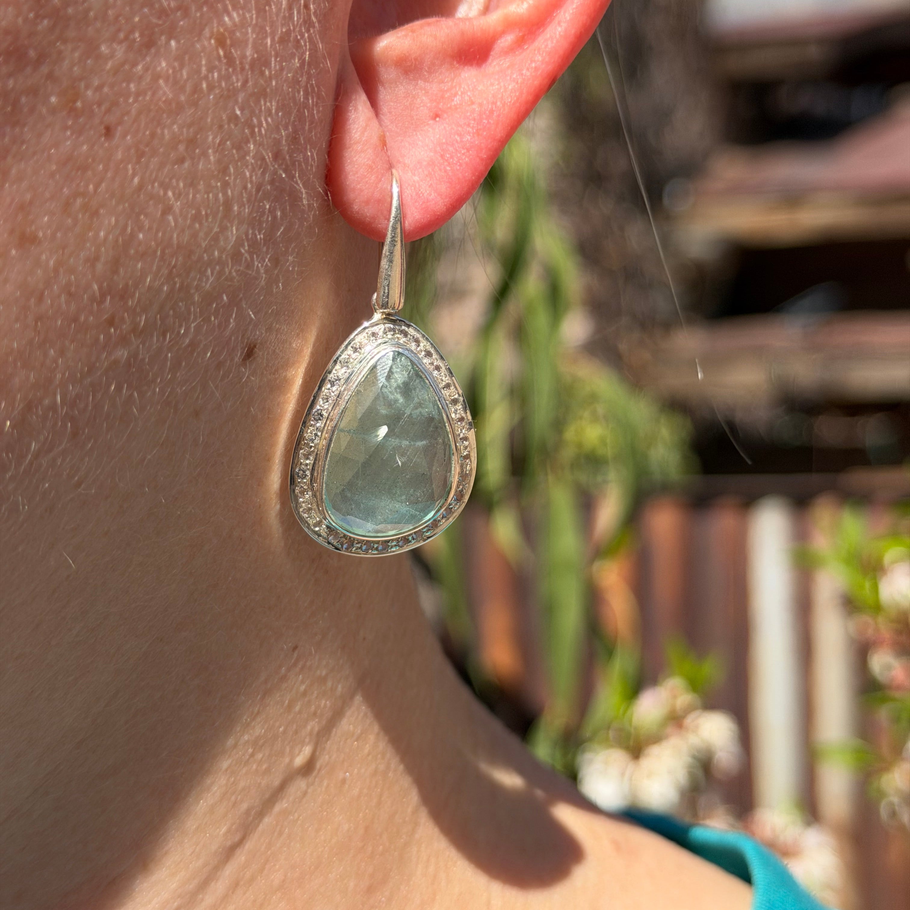 Closeup of faceted Aquamarine Dangle Earrings in Sterling Silver with white topaz halo pave worn on a model