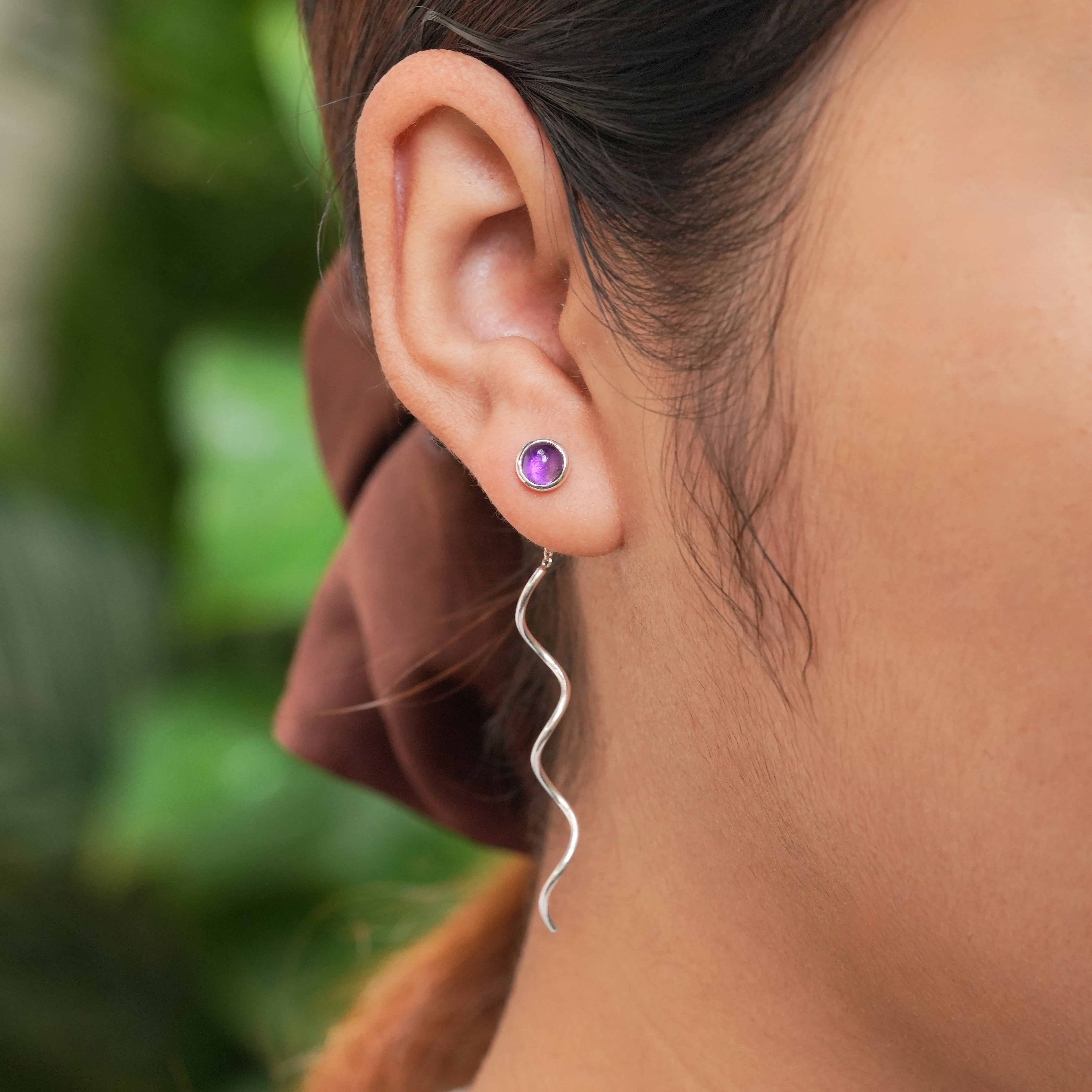 Round Amethyst stud earrings with flowing curled sterling silver wire drops worn on an ear to illustrate length and positioning. 